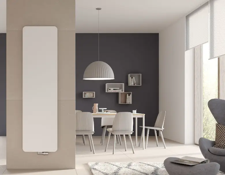 Vertikaler Kermi-Heizkörper in Weiß, montiert an einer Wand, vor einem Essbereich mit hellem Tisch und Stühlen in modernem, hellen Raum.